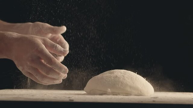 Male hands throw a piece of dough to a wooden table and sprinkle it with flour. Filmed on black background