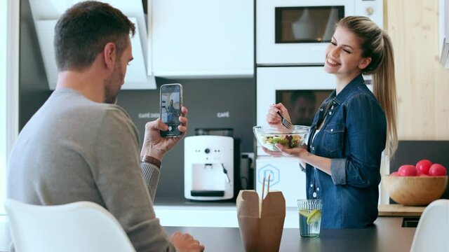 Video of funny cute couple eating and taking photos with smartphone in the kitchen at home.