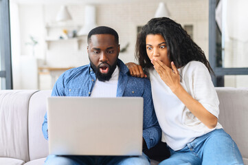 Young beautiful multiracial couple sitting on the grey sofa of new house using computer laptop...