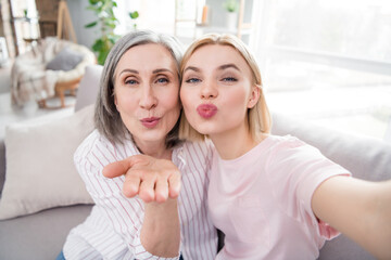 Self-portrait of two attractive elderly cheerful women sitting on cozy divan hugging sending air kiss at home house indoor
