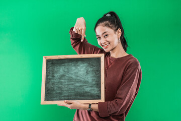 Young woman holding and pointing bottom on a blank blackboard with smiling happiness on a green background