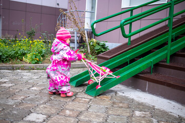 cute toddler with a toy stroller walks along steel railing ramp for wheelchair, carts and...