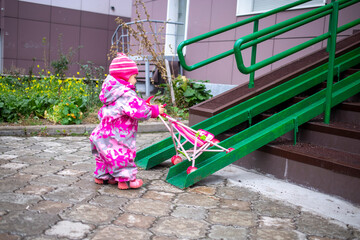 cute toddler with a toy stroller walks along steel railing ramp for wheelchair, carts and strollers. gentle descent from the stairs on the street.