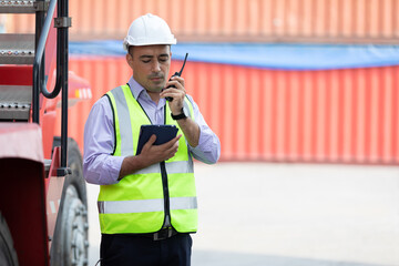 factory worker or engineer using walkie talkie and tablet for preparing a job beside truck in containers warehouse storage