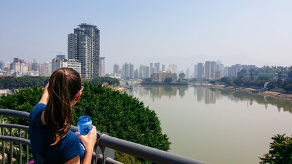 Fototapeta na wymiar A woman standing at the bridge above Min River with a panoramic view on the skyline of Fuzhou, China. The woman is pointing to the high skyscrapers. The river has brownish color. Clear and blue sky.