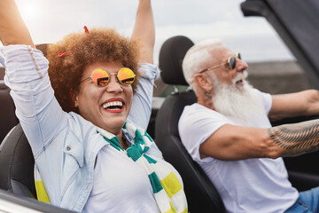 Trendy senior couple having fun inside convertible car - Multiracial mature people on a road trip...