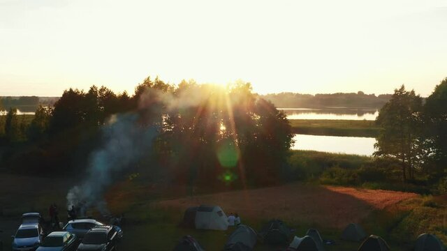Drone flyover above morning camping spot with campfire, cars and tents to large lake and epic summer sunrise panorama.