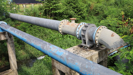 Fototapeta na wymiar Clamps and nuts to hold old pipes on posts. A blue main water supply pipe with clamps and bolts connecting pipes on a concrete column base across the water canal with green plants. Selective focus