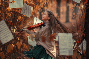 A beautiful girl plays the violin among the autumn orange foliage in the forest and with music sheets in the background. © Kiryakova Anna