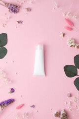 White tube of cosmetic cream with flowers and green leaves on pink background. Flat lay
