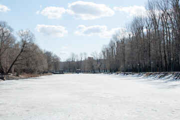 Winter panorama of a frozen lake, trees and poles with lanterns in the park.