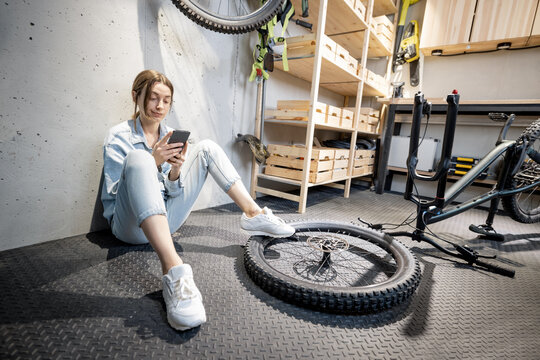 Young relaxed handywoman sitting with phone while repairing her bicycle in the well equipped workshop at home. DIY concept