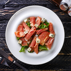 Salad of ham, red tomato, lettuce leaves, cheese in a white, round plate on a black, textured wood background