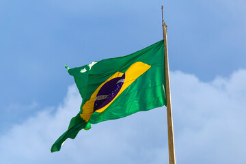 Bandeira do Brasil com nuvens e céu azul ao fundo.