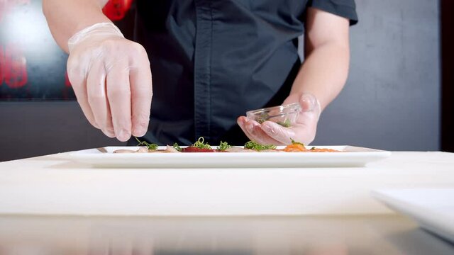 Close-up of male chef hands in gloves preparing delicious appetizer of fresh fish and squid with truffle sauce and herbs on a plate.