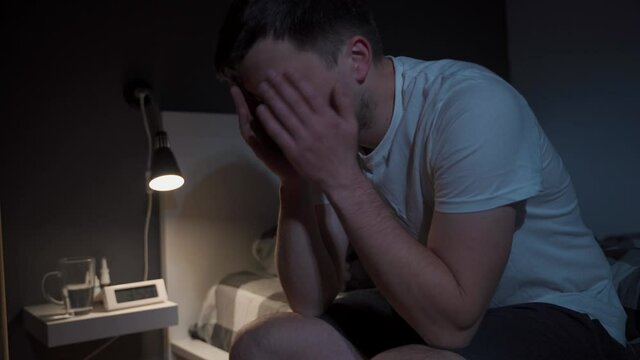 Handsome young man suffering from headache while sitting on bed at night. Exhausted man sitting on bed with hand on head. Male depressed and upset in bedroom. Guy constantly struggles with insomnia