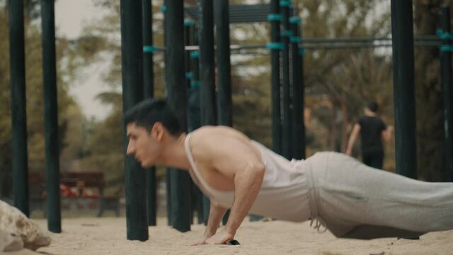hombre haciendo flexiones en el parque, hombre de pelo negro haciendo deporte al aire libre, hombre musculoso en camiseta de tirantes  blanca haciendo ejercicio