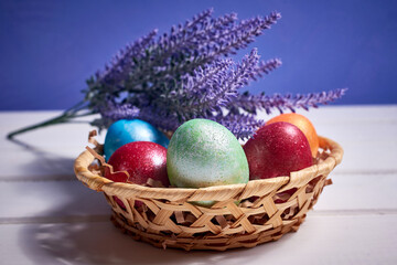 A small basket with colorful painted eggs and a bouquet of lavender on a white wooden background. Orthodox Easter composition.