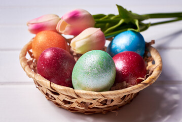 A small basket with colorful painted eggs and a bouquet of tulips on a white wooden background. Orthodox Easter composition.
