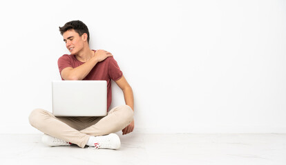Teenager man sitting on the flor with his laptop suffering from pain in shoulder for having made an effort
