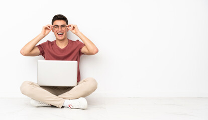 Teenager man sitting on the flor with his laptop with glasses and surprised