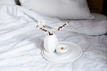 Blooming apricot tree branches and burning candle on round tray in bedroom interior, white spring home decoration