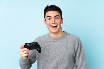 Teenager caucasian man playing with a video game controller isolated on blue background with surprise and shocked facial expression