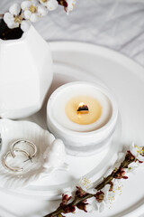 Blooming apricot tree branches and burning candle on round tray in bedroom interior, white spring home decoration