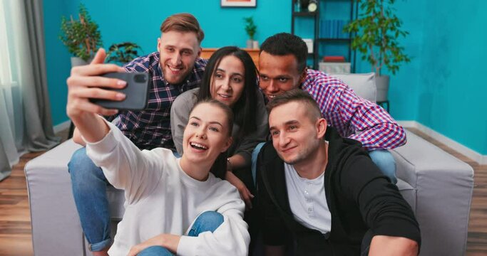Joyous multi-ethnic group of friends sitting on couch together and taking selfie with smartphone while watching soccer match on TV