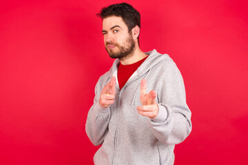 Joyful Young caucasian man wearing tracksuit over red background wink and points index fingers at...