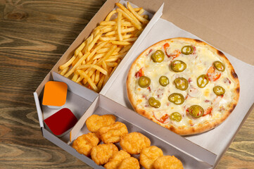 Crispy pizza with squid rings, pickles, fresh tomato and cheese in a set menu box with golden fries, nuggets and cheese and tomato sauces on a wooden table top