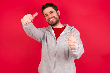 Young caucasian man wearing tracksuit over red background approving doing positive gesture with hand, thumbs up smiling and happy for success. Winner gesture.