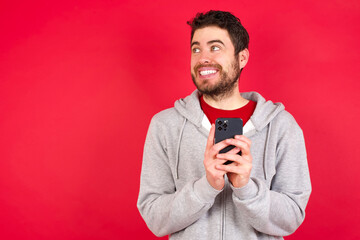 Young caucasian man wearing tracksuit over red background holding a smartphone and looking sideways at blank copyspace.