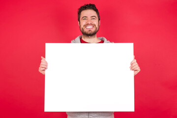 Happy Young caucasian man wearing tracksuit over red background Holding Empty Paper Board Advertising Offer Text Standing. Autumn Advertisement Banner.