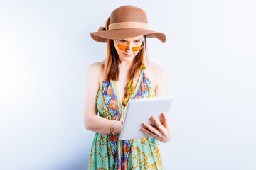 beautiful young woman in summer dress, hat and yellow glasses on white background with copy space checking the tablet from work. concept take work on vacation