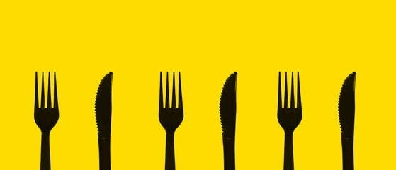 Black knife and fork, plastic utensil for takeaway dining laying vertical alignment on yellow table. Monotone color object repeating on background, isolated. Minimlist pattern concept with copy space.