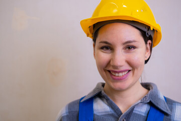 A beautiful construction worker girl in a yellow hard hat and a checkered shirt looks at the camera, smiles and winks against the background of a stucco wall.