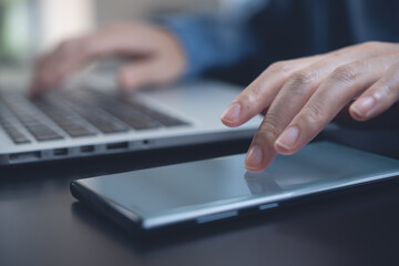 Closeup of woman finger touching on mobile phone screen and working on laptop computer on office table