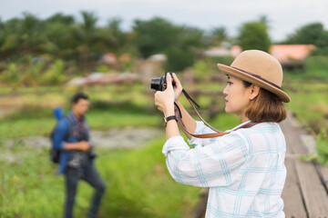 A tourist woman enjoy taking photo of landscape during her summer vacation holiday in the greenery field. A stylish traveler woman in hat taking photo of herself and landscape while relaxing