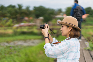 A tourist woman enjoy taking photo of landscape during her summer vacation holiday in the greenery field. A stylish traveler woman in hat taking photo of herself and landscape while relaxing