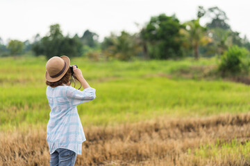 A tourist woman enjoy taking photo of landscape during her summer vacation holiday in the greenery field. A stylish traveler woman in hat taking photo of herself and landscape while relaxing