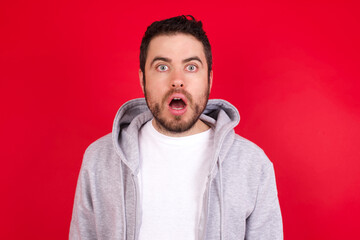 young handsome caucasian man in sports clothes against red wall having stunned and shocked look, with mouth open and jaw dropped exclaiming: Wow, I can't believe this. Surprise and shock