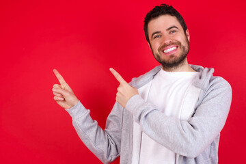 young handsome caucasian man in sports clothes against red wall with positive expression, indicates with fore finger at blank copy space for your promotional text or advertisement.