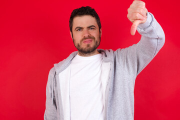 Discontent young handsome caucasian man in sports clothes against red wall shows disapproval sign, keeps thumb down, expresses dislike, frowns face in discontent. Negative feelings.