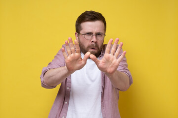 Frightened Caucasian man with glasses makes a stop gesture, he puts out palms and looks anxiously. Yellow background.
