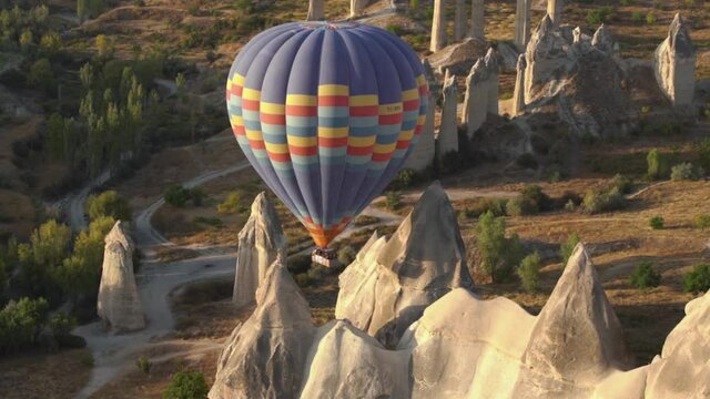 Large hot air balloon with coloured stripes flies over brown bare rocky cliffs and stone pillars among rare bushes and ground roads under bright, sunlight.