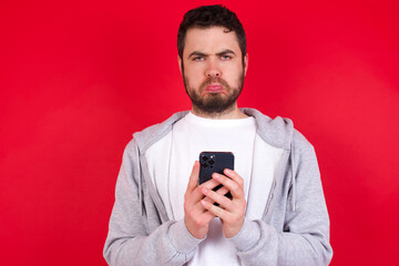 Portrait of a confused young handsome caucasian man in sports clothes against red wall holding mobile phone and shrugging shoulders and frowning face.
