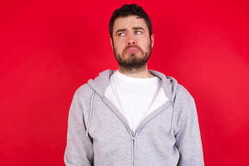 Dissatisfied young handsome caucasian man in sports clothes against red wall purses lips and has unhappy expression looks away stands offended. Depressed frustrated model.