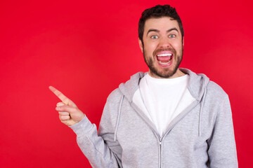 young handsome caucasian man in sports clothes against red wall points aside on copy blank space. People promotion and advertising concept