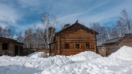 A country house made of unpainted natural logs stands among the snowdrifts. There are shutters on the small windows. Background - blue sky, white clouds, bare branches and tree trunks.